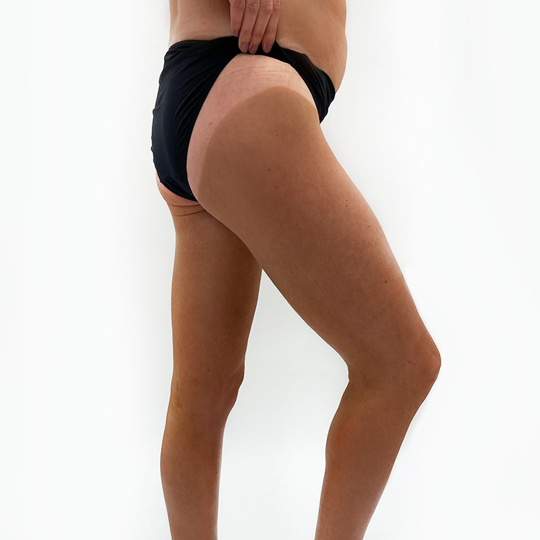 Image of a woman in a black bikini. The woman has just had a spray tan and she is lifting the side of her bikini up to show the difference the colour of her skin after a dark spray tan by He-Shi
