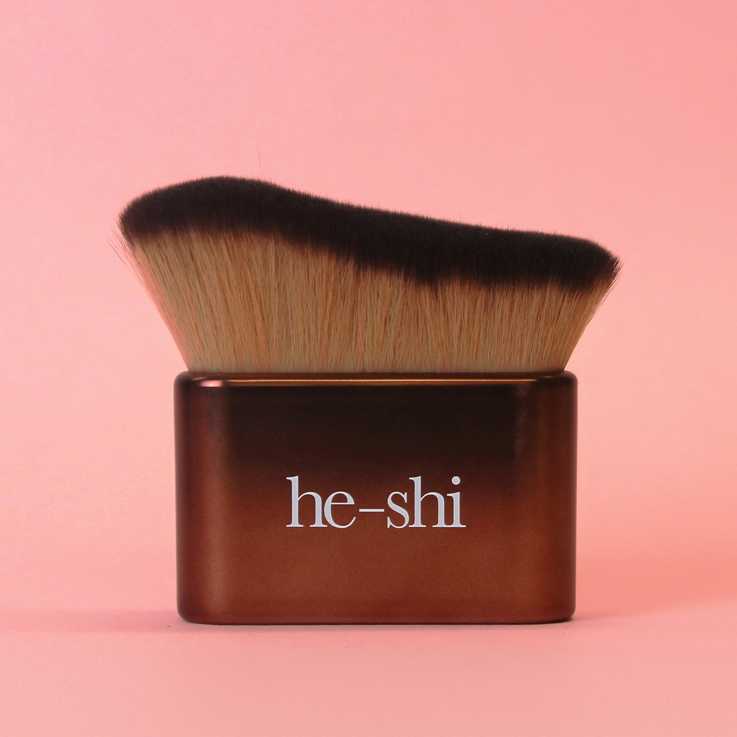 He-Shi Tanning brush with brown handle and black bristles on a pink background, featuring the brand name 'he-shi'.
