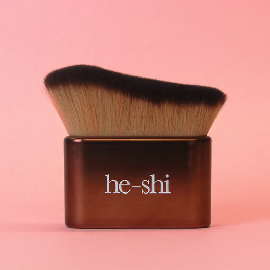He-Shi Tanning brush with brown handle and black bristles on a pink background, featuring the brand name 'he-shi'.